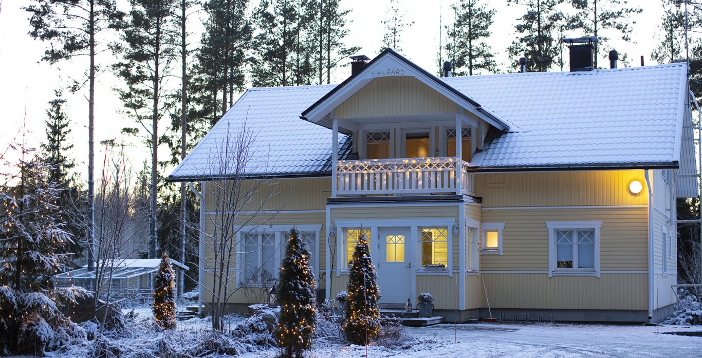 Keltainen omakotitalo talvella. Etupihan puissa ja parvekkeella jouluvalot.
