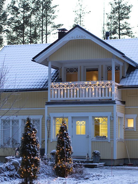 Keltainen omakotitalo talvella. Etupihan puissa ja parvekkeella jouluvalot.
